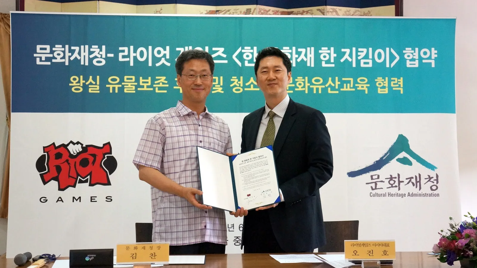 Two men holding a signed agreement between Riot Games and Korea's Cultural Heritage Administration, in front of a partnership backdrop