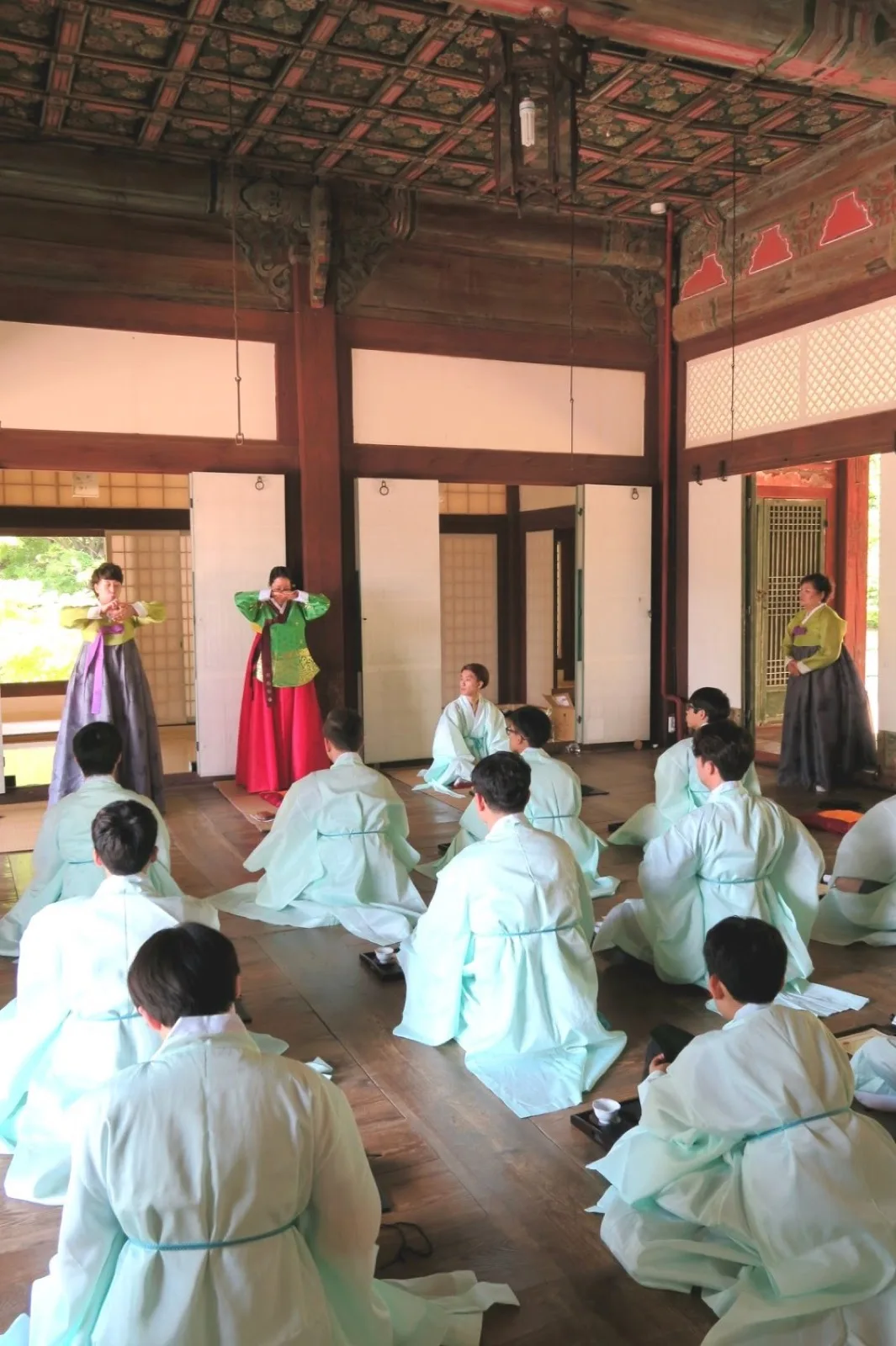 Students in light-colored traditional hanbok seated on the floor of a historic hall, with instructors in colorful hanbok demonstrating