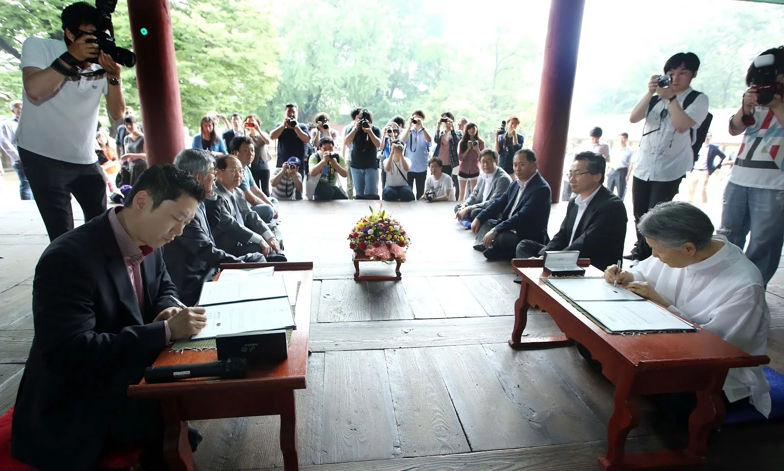 Signing ceremony on a wooden pavilion floor, with officials seated at low red tables and photographers recording