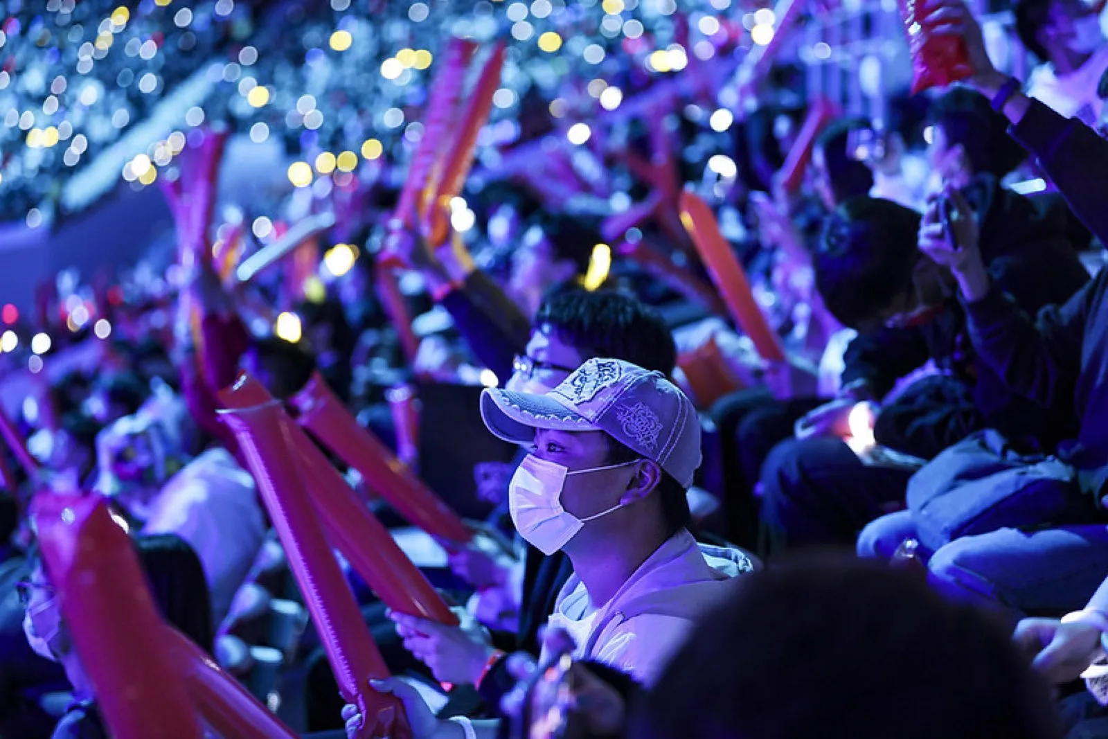 Close-up of a masked fan in a cap among a crowd waving glowing thunder sticks at Worlds 2020